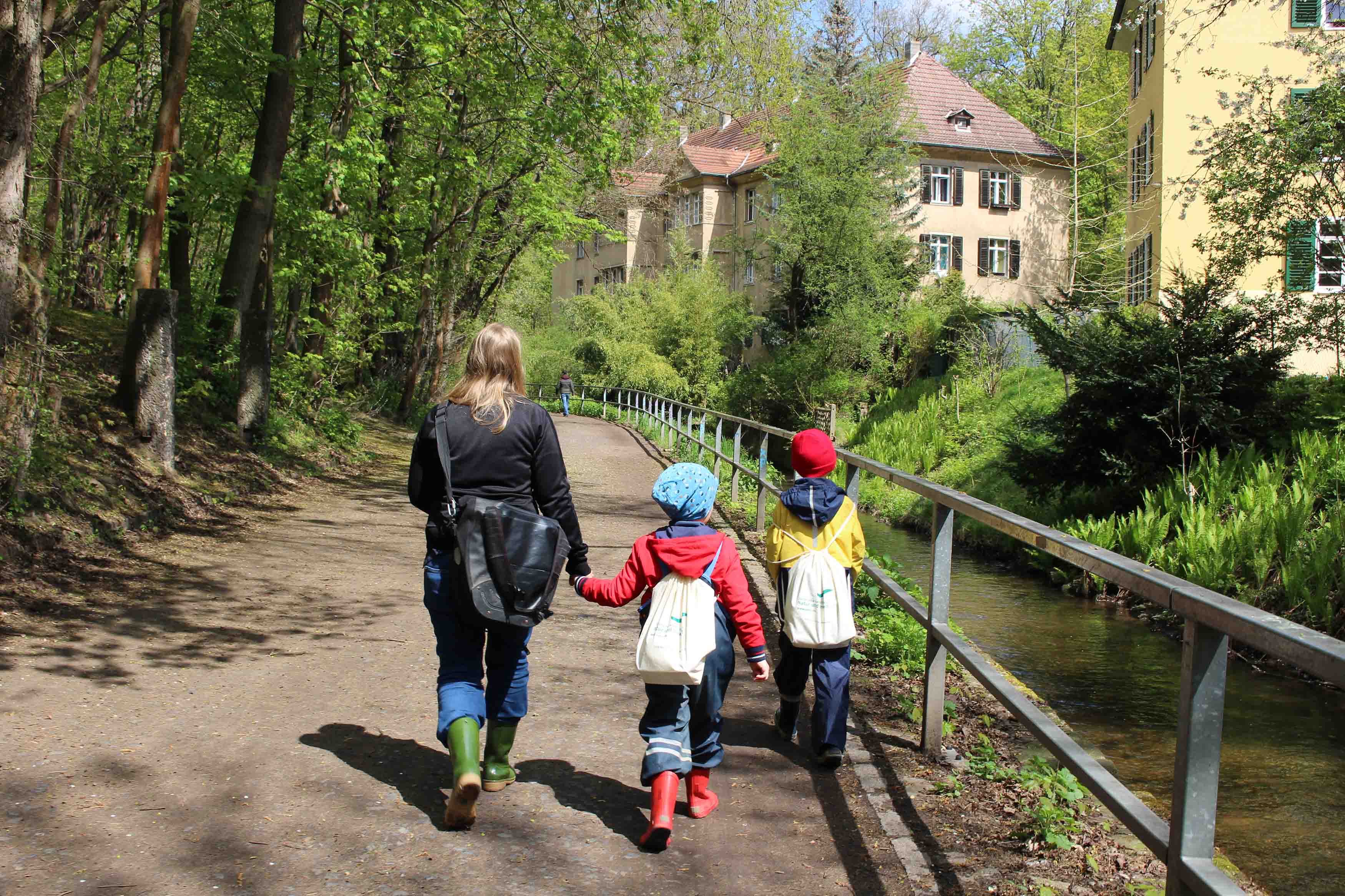 KI generiert: Das Bild zeigt eine erwachsene Person und zwei Kinder, die auf einem Weg entlang eines Baches spazieren gehen. Im Hintergrund sind Gebäude und üppige Vegetation zu sehen.