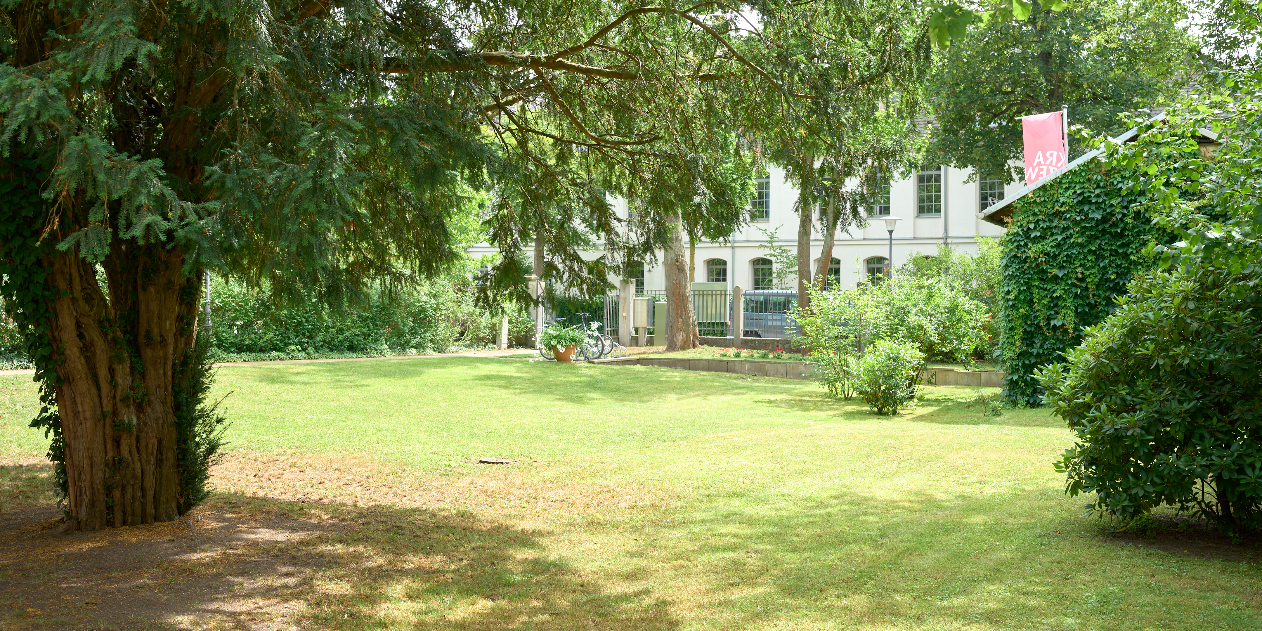 KI generiert: Das Bild zeigt einen gepflegten Garten mit Rasenflächen, Sträuchern und einem großen Baum im Vordergrund. Im Hintergrund ist ein weißes Gebäude mit mehreren Fenstern und einem rosa Schild zu sehen.