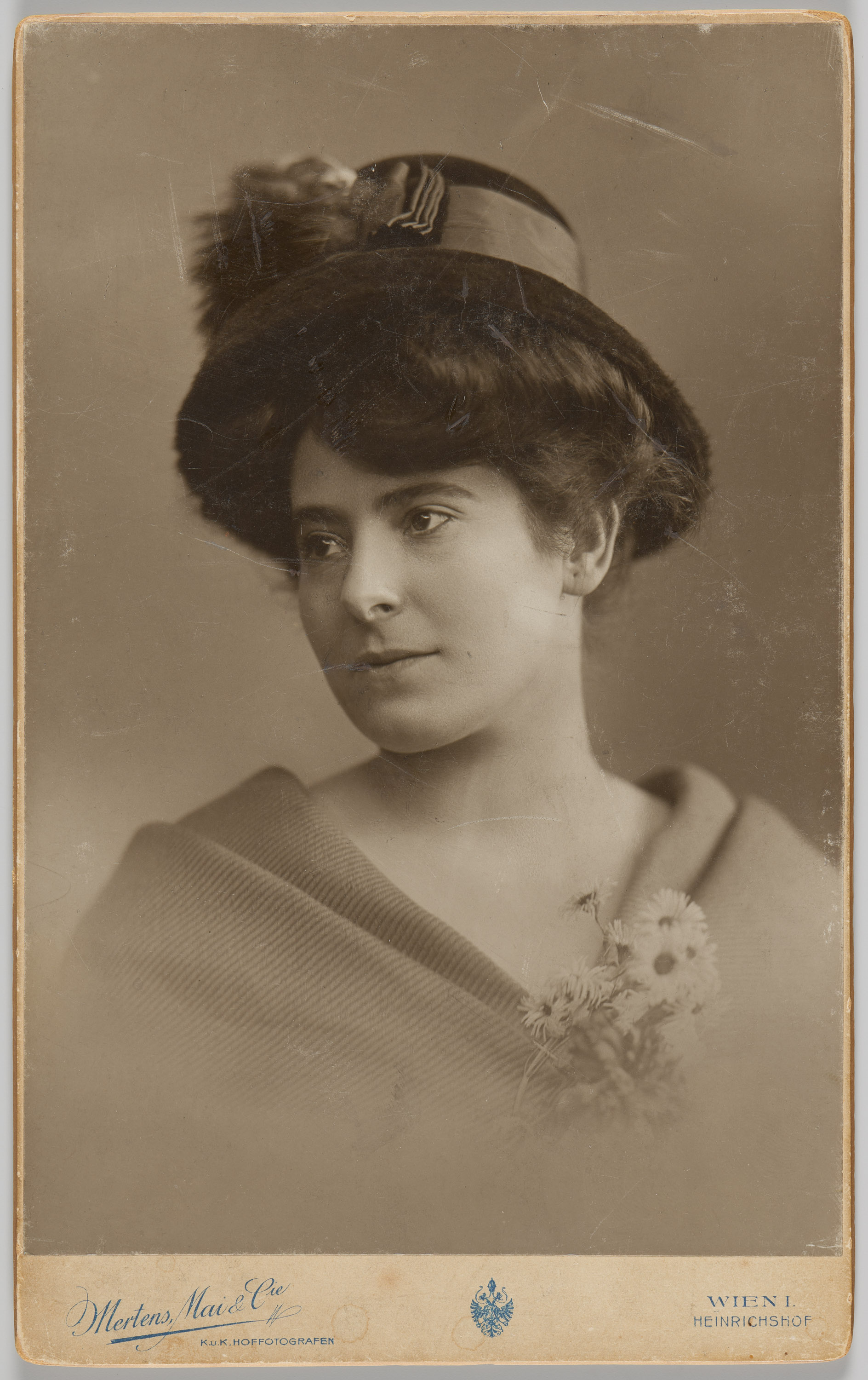 KI generiert: Das Bild zeigt eine Frau in einem historischen Porträt, gekleidet mit einem Hut und einem Umhang oder Schal, während sie zur Seite schaut. Sie hält einen kleinen Blumenstrauß in der Hand.