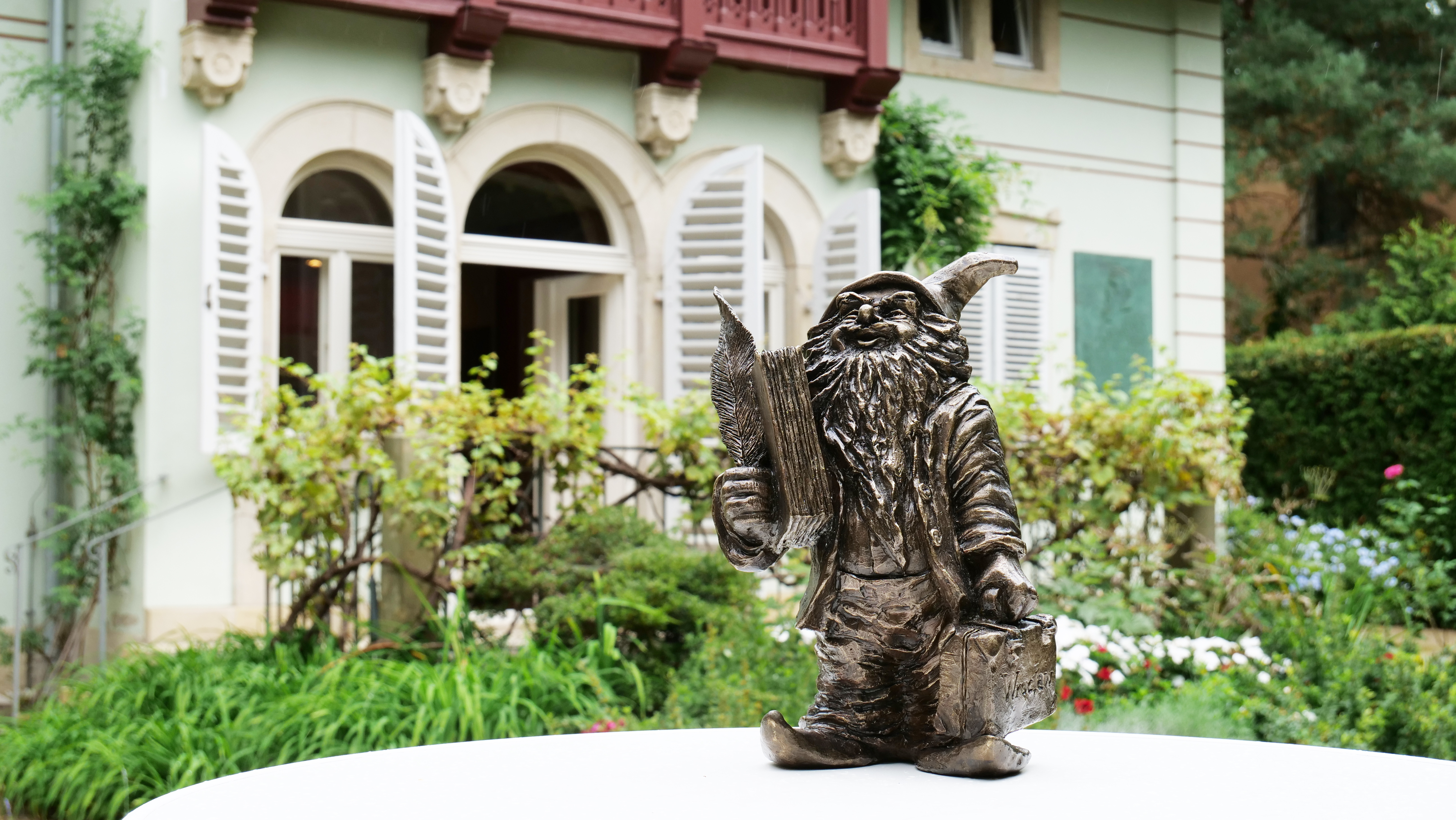 KI generiert: Das Bild zeigt eine Statue eines Gartenzwergs, der eine Zeitung in einer Hand und eine Tasche in der anderen hält. Im Hintergrund ist ein Haus mit geöffneten Fensterläden und einem üppigen Garten zu sehen.