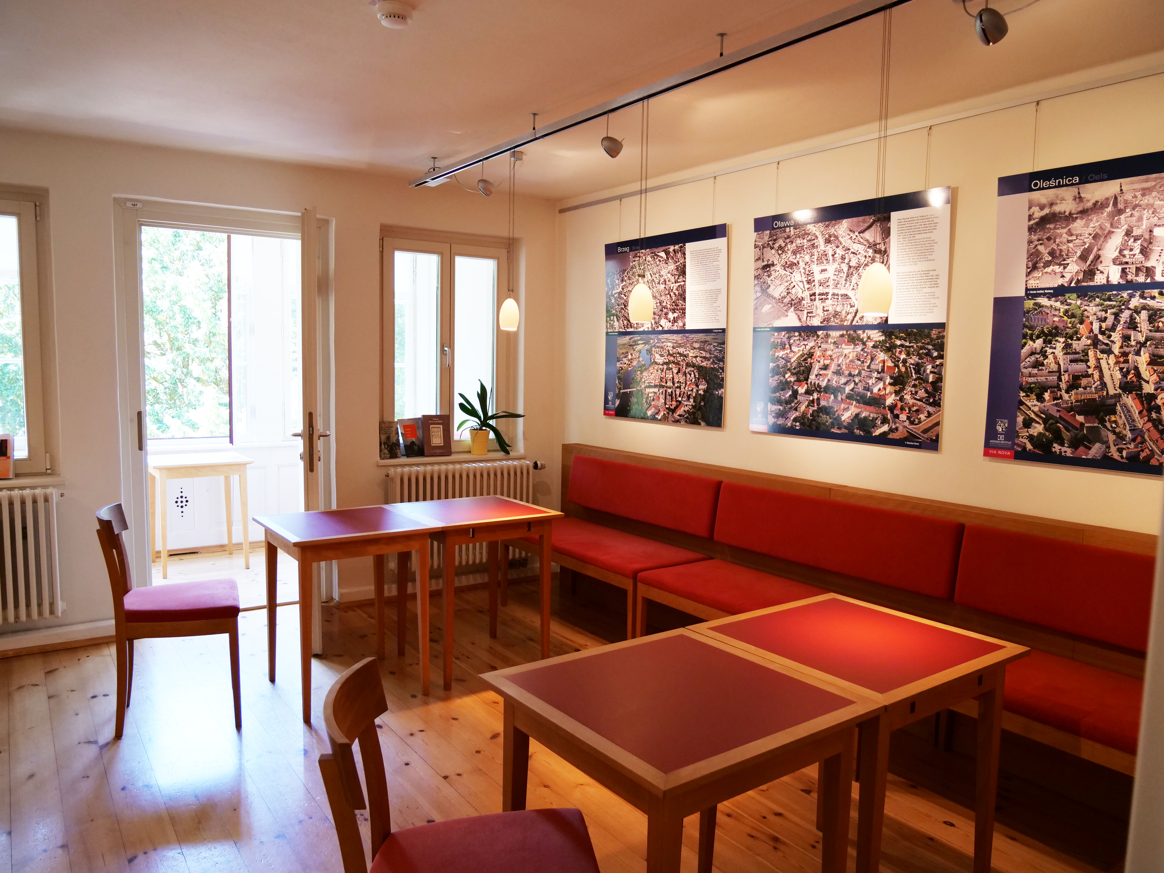 KI generiert: Das Bild zeigt einen gemütlichen Raum mit Holzboden und rotem Mobiliar. An den Wänden hängen Stadtpläne und Bilder von Stadtansichten, die eine entspannte Atmosphäre schaffen.