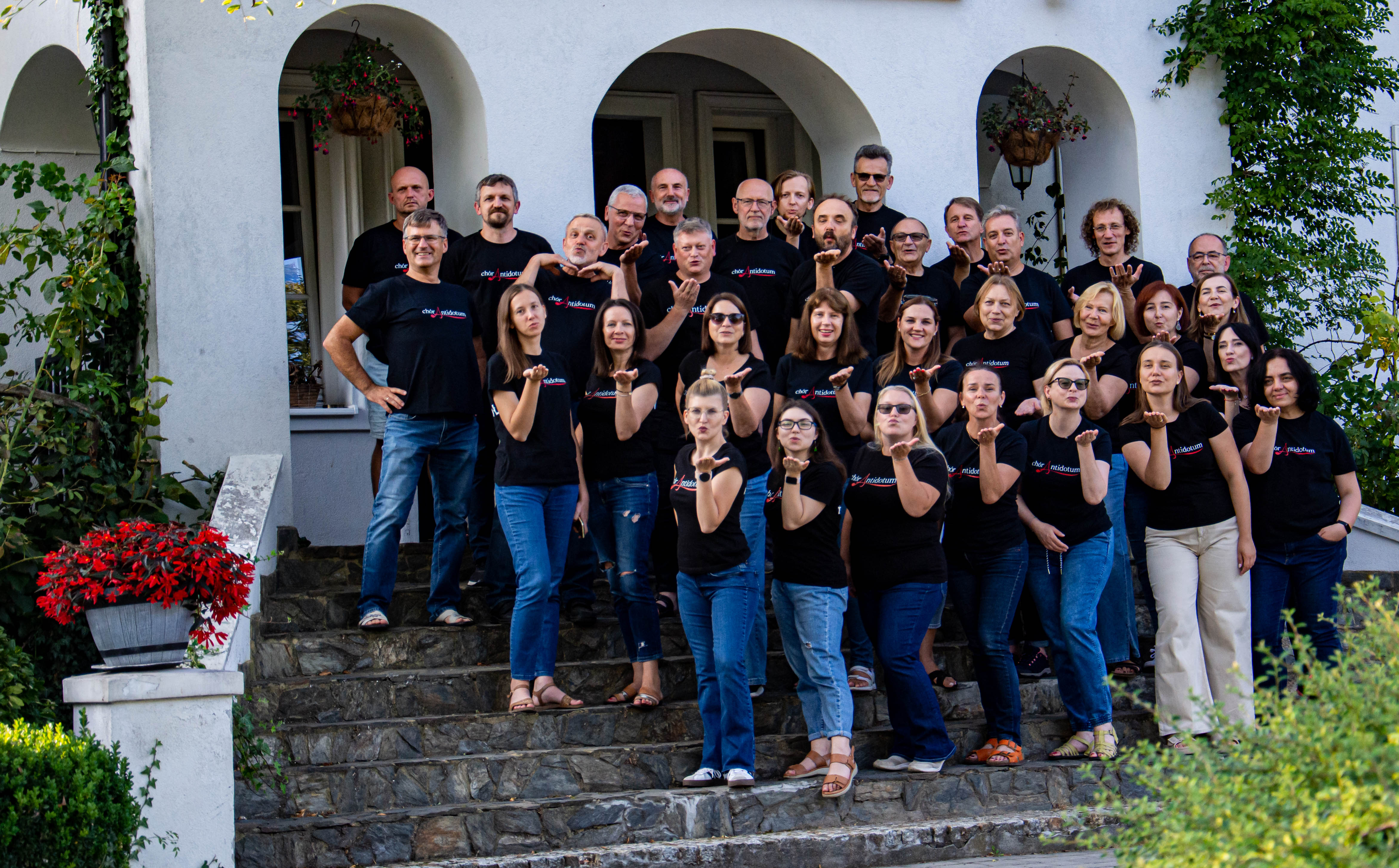KI generiert: Eine Gruppe von Menschen posiert auf einer Treppe vor einem Gebäude und trägt dabei einheitliche schwarze T-Shirts. Sie scheinen in guter Stimmung zu sein und machen eine Handgeste.