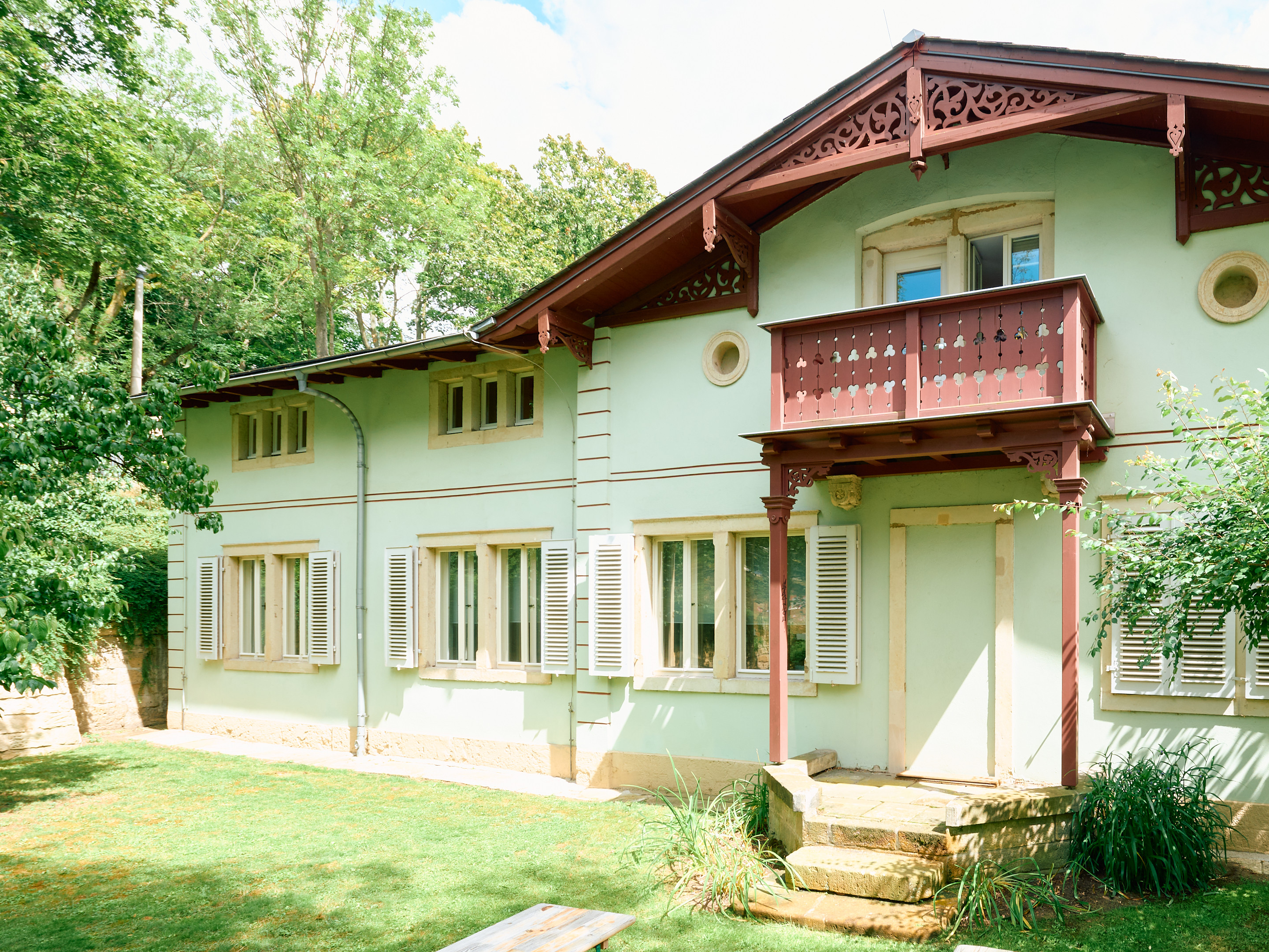 KI generiert: Das Bild zeigt ein hellgrünes Haus mit einer kleinen Veranda und weißen Fensterläden, das von Bäumen und Grünflächen umgeben ist. Die Architektur des Hauses wirkt gemütlich und einladend.