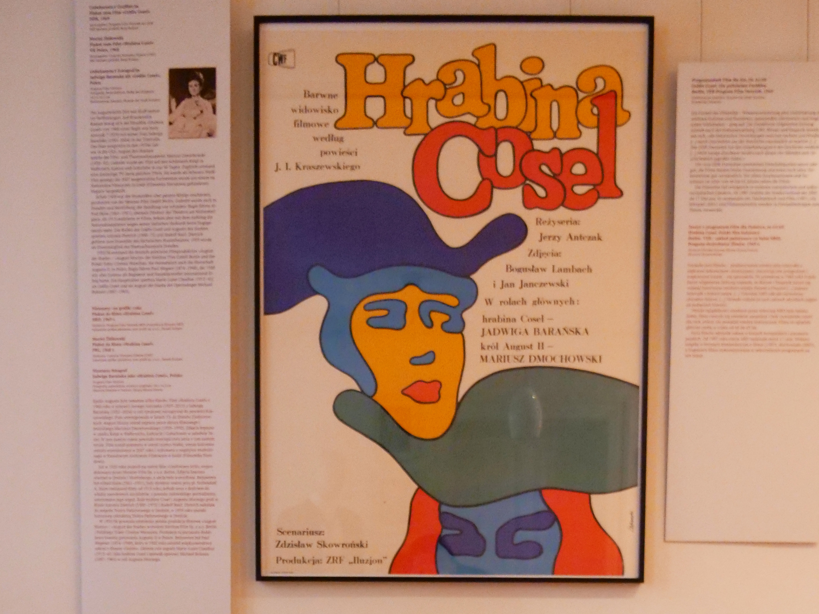 AI generated: The image shows a colourful poster with the title "Hrabina Cosel" on a wall in the background, flanked by two text panels. In the foreground, a magazine with the title "NBI" lies on a table.