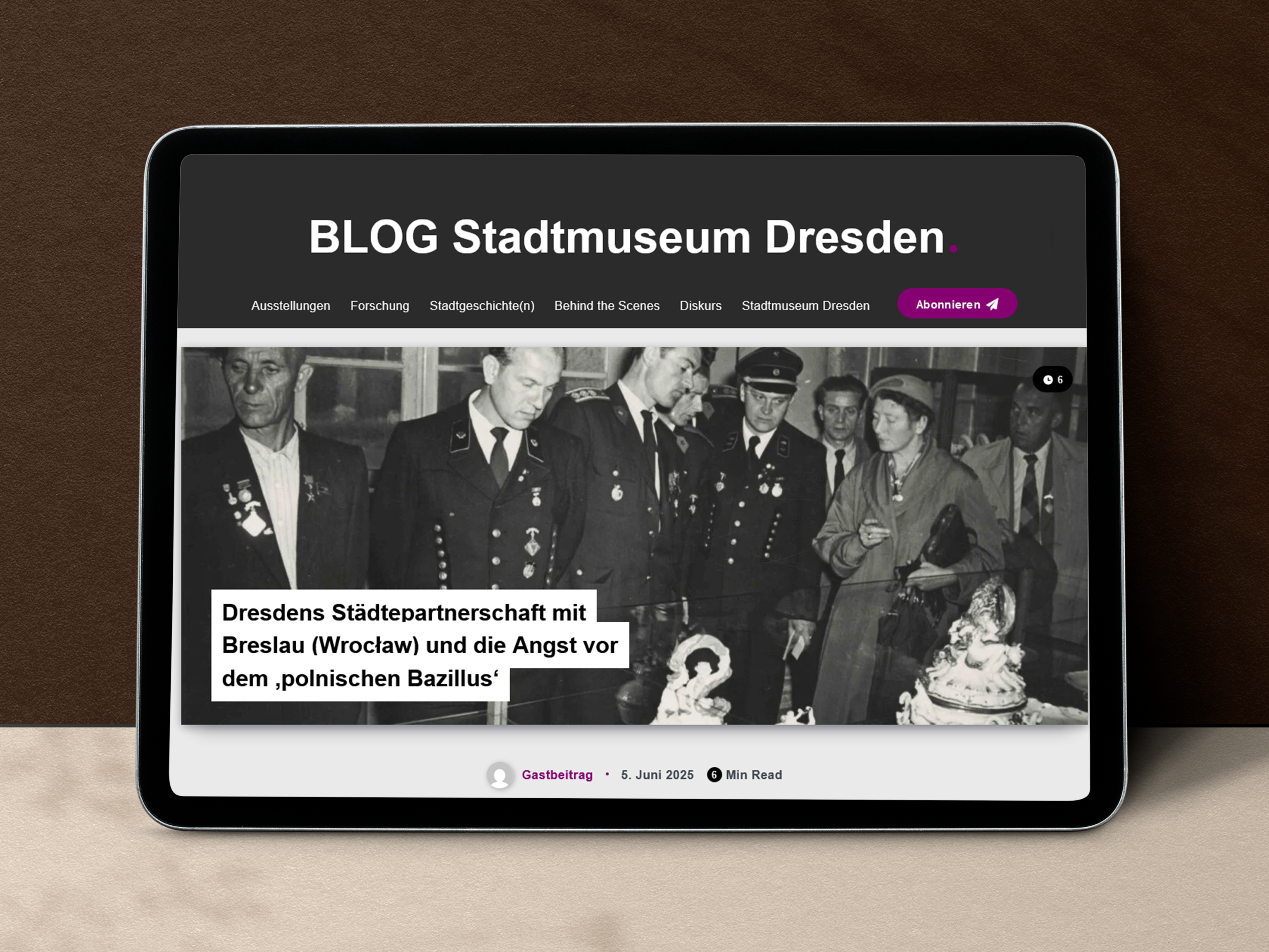 KI generiert: Ein Tablet zeigt die Webseite des "BLOG Stadtmuseum Dresden" mit einem Artikel über die Städtepartnerschaft Dresdens mit Breslau (Wrocław). Das Bild im Hintergrund zeigt eine historische Szene mit mehreren Personen in Uniform.